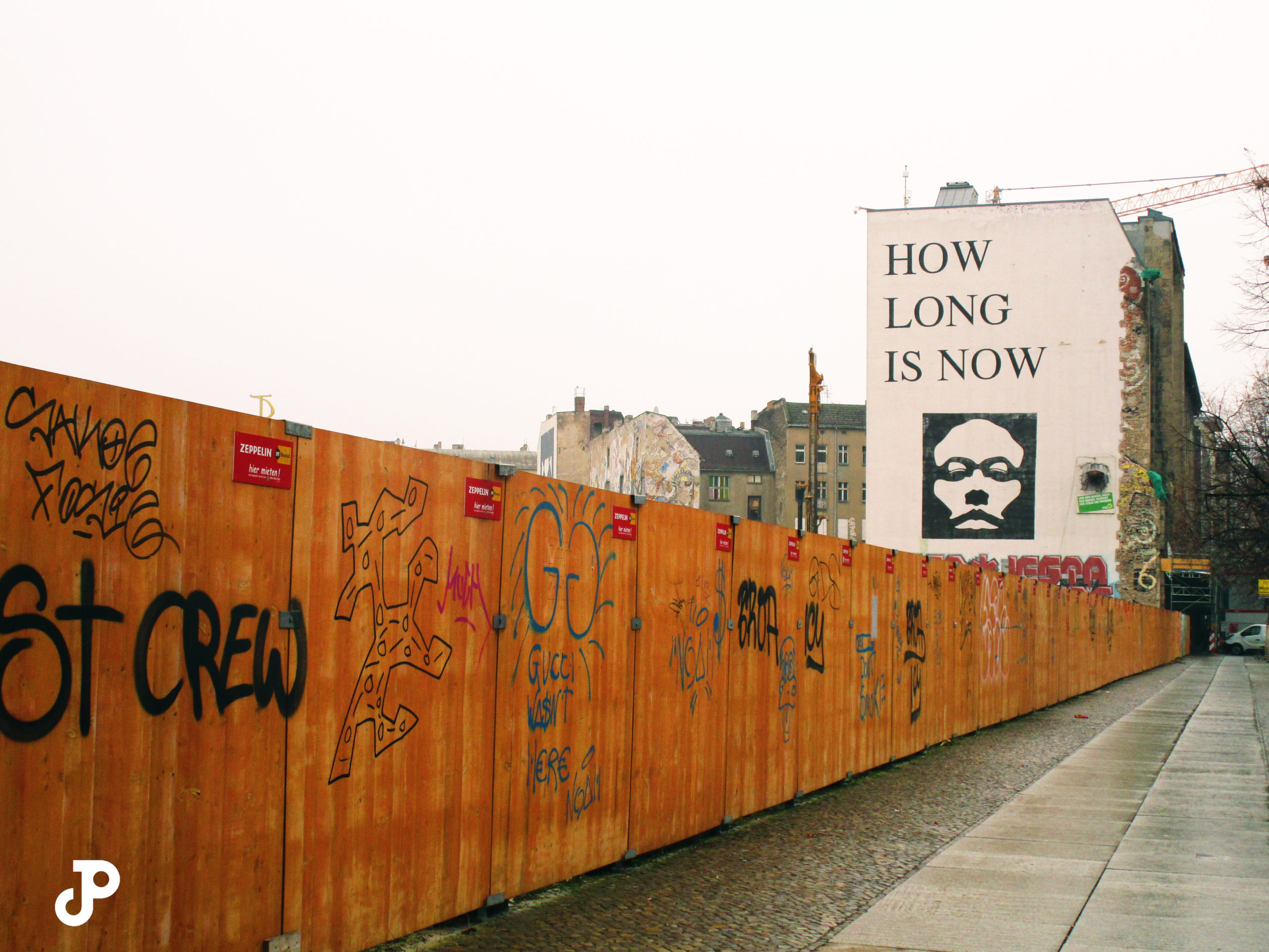 a wooden fence covered in graffiti leading to a building in the distance, which is painted with a mural that reads, how soon is now?