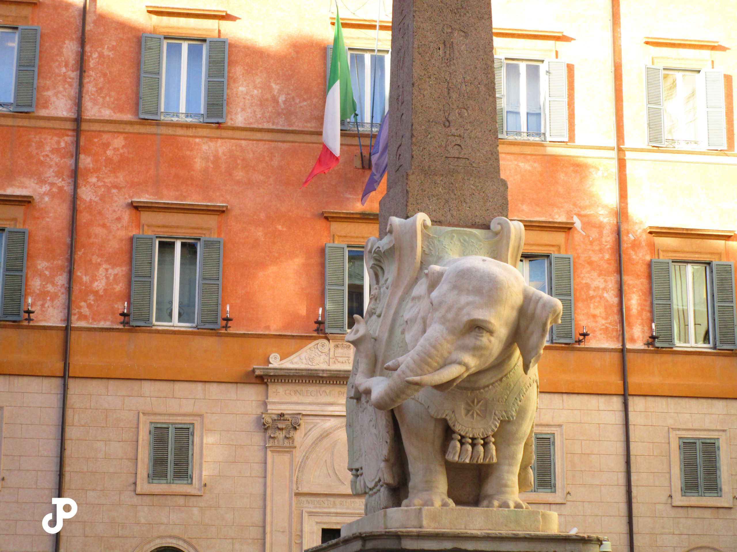 a marble elephant statue in the foreground, and in the background an orange building with an Italian flag