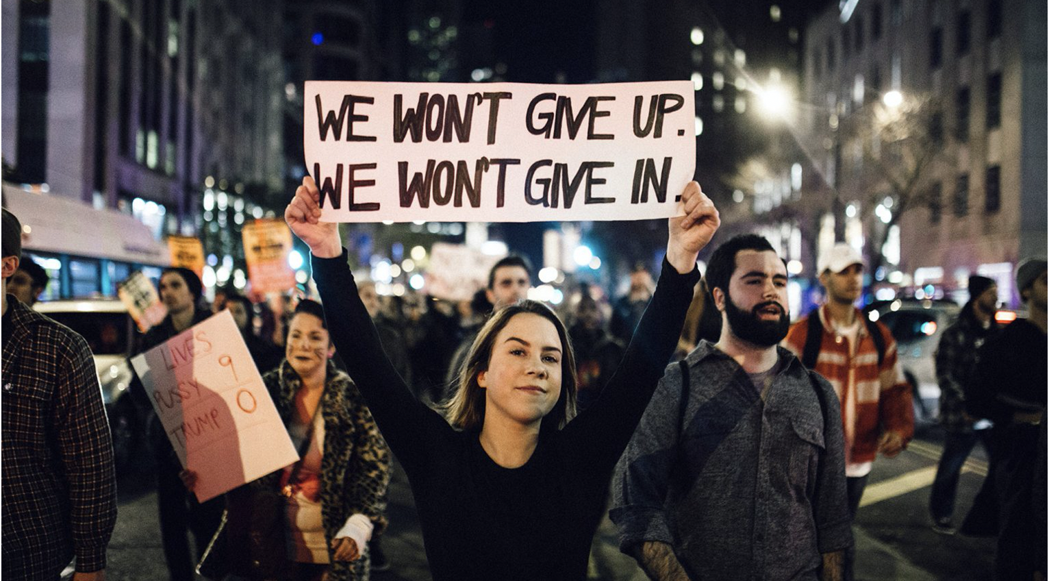 People at a protest, with a woman in the foreground holding a sign that reads, we won't give up, we won't give in