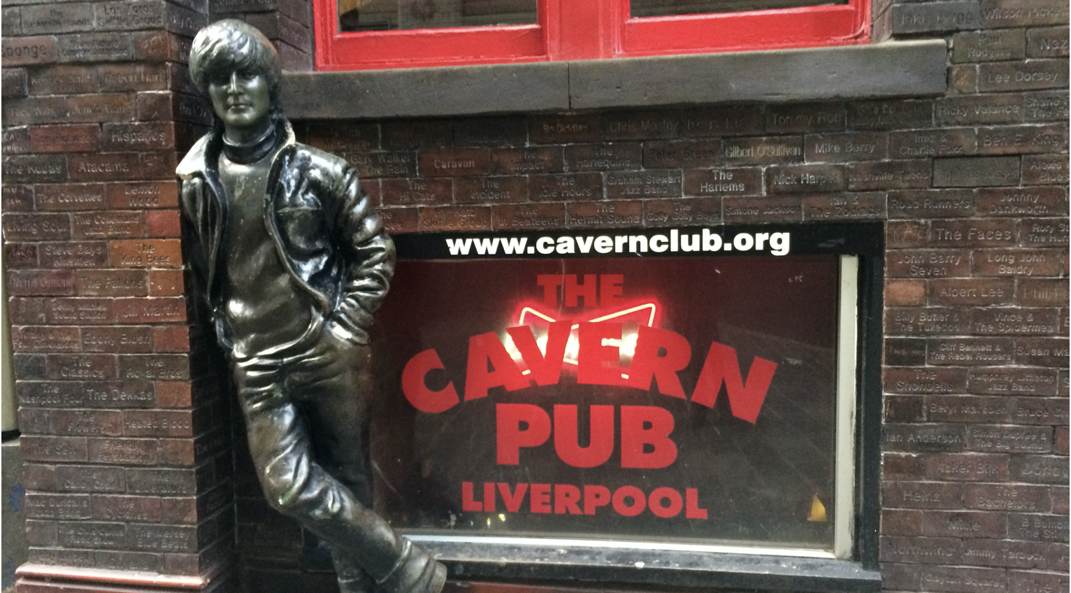 A statue of John Lennon standing beside a glass window with red painted letters that read The Cavern Plub Liverpool